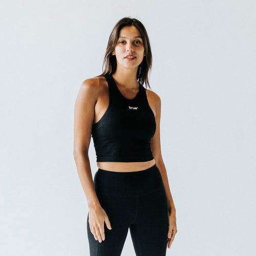 Female model in studio wearing black ribbed racerback crop tank top with small "true" logo and high-waist black leggings — women's activewear crop top for yoga, gym, athleisure.