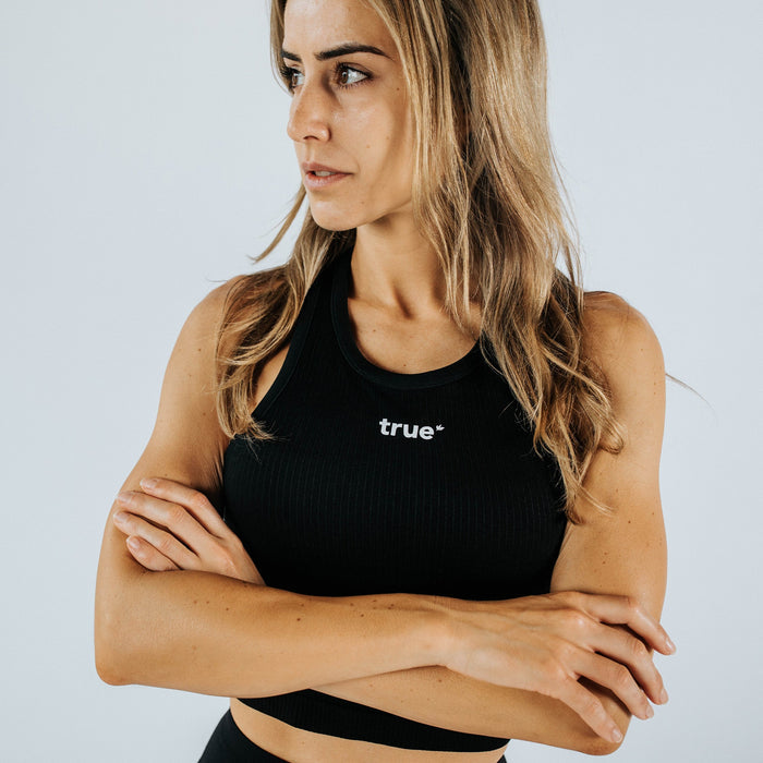 Woman wearing black ribbed women's crop tank top with white "true" logo, arms crossed, athletic fitness model in studio on light background — close-up product shot for activewear.