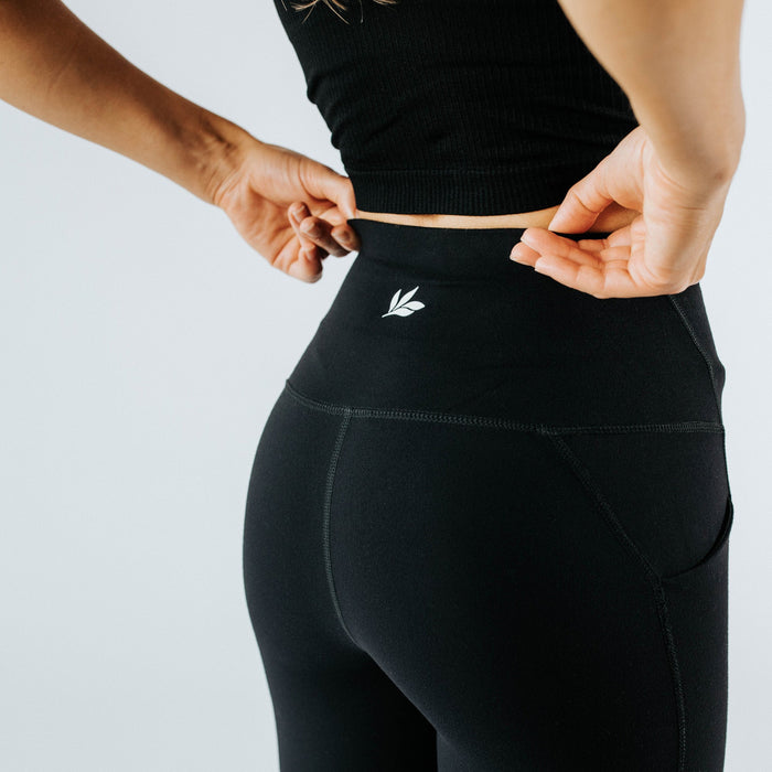 Close-up back view of women's high‑waist black active leggings with side pocket and small logo, model adjusting waistband — seamless squat‑proof workout leggings for yoga, running and gym.