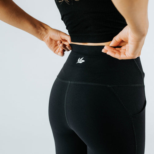 Close-up back view of women's high‑waist black active leggings with side pocket and small logo, model adjusting waistband — seamless squat‑proof workout leggings for yoga, running and gym.