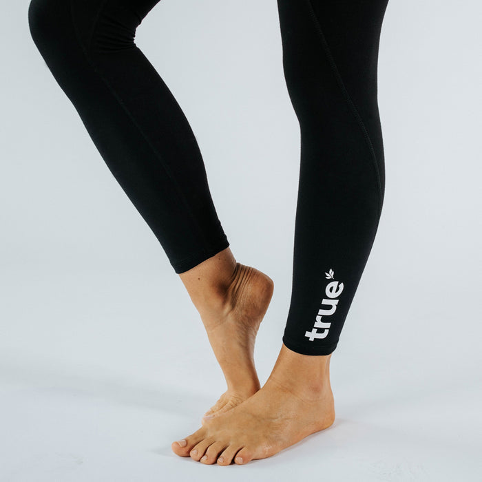 Black women's active leggings with white "true" logo on lower calf, barefoot model in yoga pose on white studio background, close-up of legs and ankles.