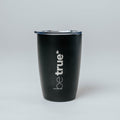 Black insulated stainless-steel travel coffee tumbler with clear lid and vertical "be true" logo, product shot on light gray background — reusable travel mug for coffee and tea.