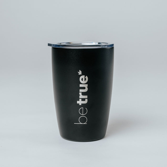 Black insulated stainless-steel travel coffee tumbler with clear lid and vertical "be true" logo, product shot on light gray background — reusable travel mug for coffee and tea.