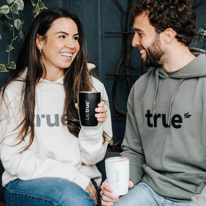 Two people smiling on an outdoor cafe bench, wearing "true" logo hoodies and holding black and white insulated "be true" travel coffee cups, lifestyle shot for sustainable coffee tumbler branding.