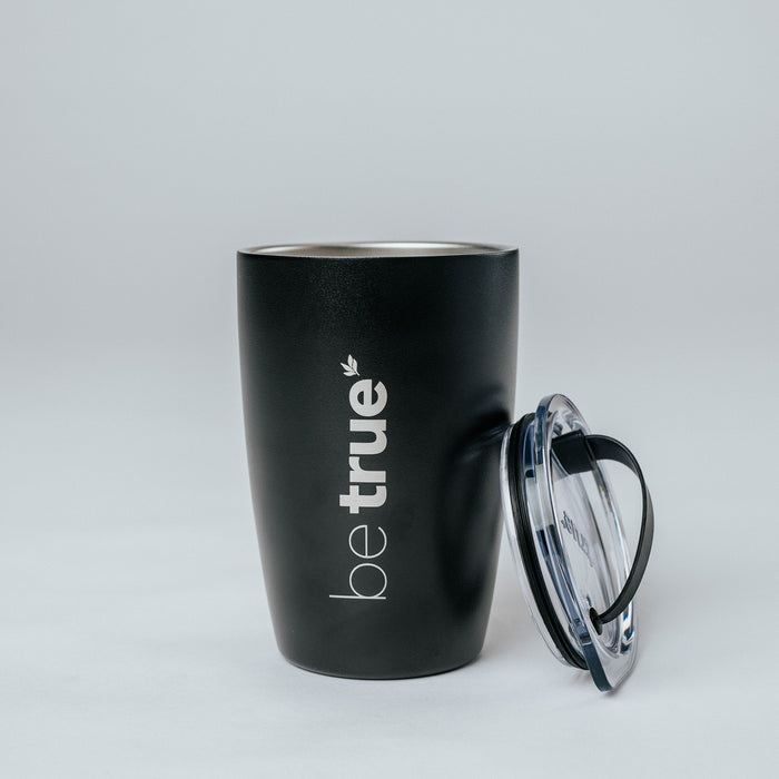Black insulated stainless steel travel mug with clear plastic lid beside it, matte black finish and vertical white "be true" logo, on a neutral studio background.