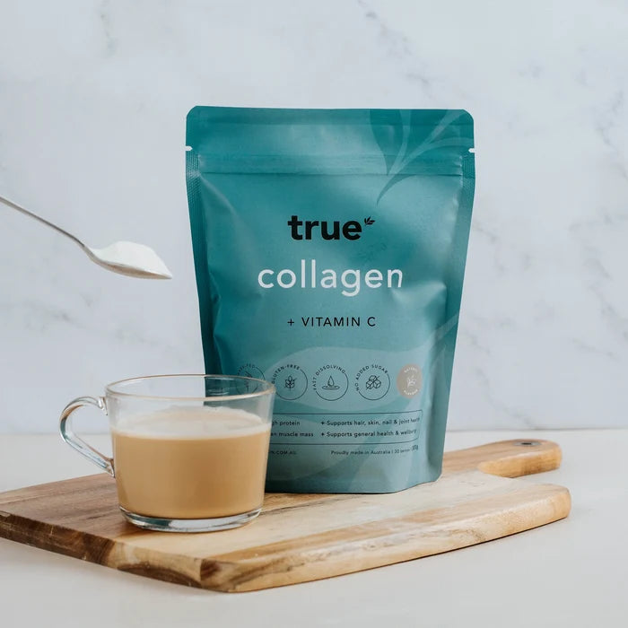 True Collagen + Vitamin C powder pouch (teal packaging) beside a glass cup of mixed collagen latte on a wooden board, spoon sprinkling powder — collagen supplement made in Australia, beauty and joint support.