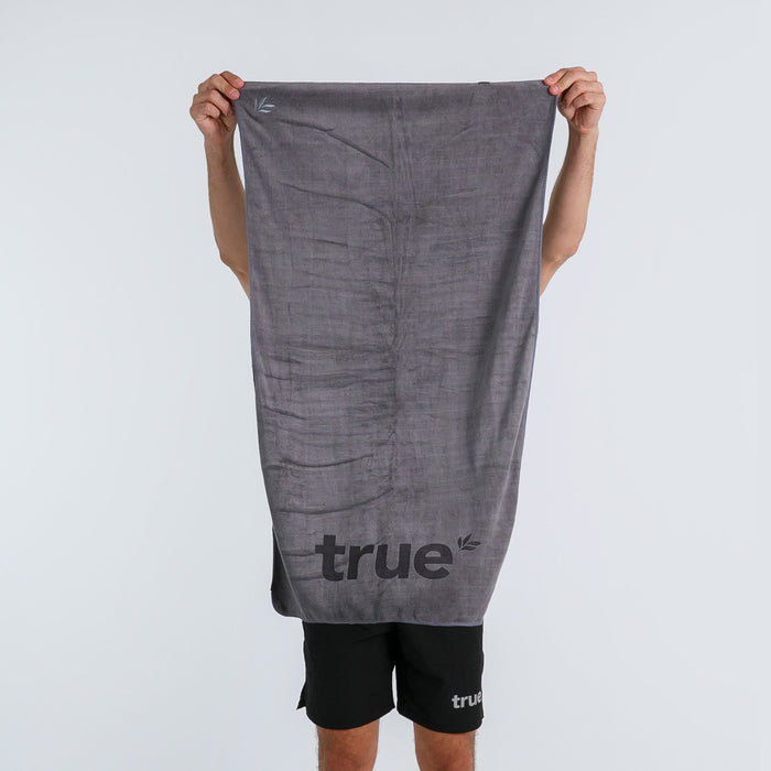 Grey True microfiber gym towel with black "true" logo held up by person against a white studio background — quick-dry fitness workout towel for gym, sports and travel.