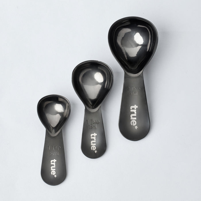 True brand matte black stainless steel 3-piece measuring spoon set — scoop style spoons 1 tsp (5 ml), 1 tbsp (15 ml), 2 tbsp (30 ml) on light gray background, for baking and cooking.