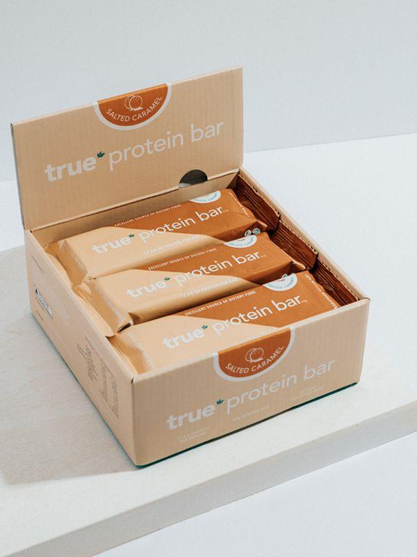 Open cardboard box of True Protein Bar salted caramel flavor showing four individually wrapped protein bars on a white countertop—healthy snack, fitness protein bar product image