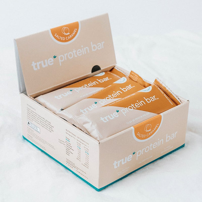 Box of True Protein Salted Caramel protein bars — beige and caramel-wrapped nutrition bars in open retail carton on white background.