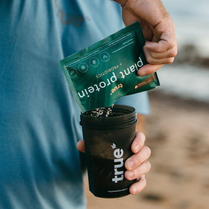 Man pouring green True Plant Protein sachet into black True shaker cup on a beach background
