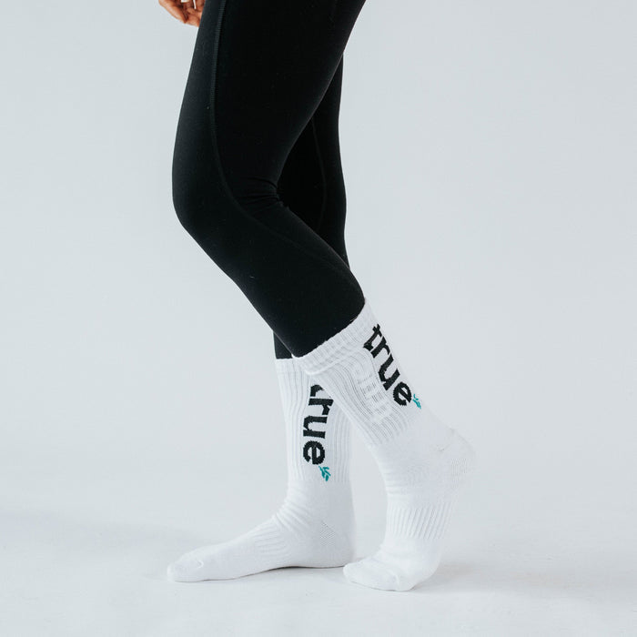 Product photo of a model wearing white crew athletic socks with black "true" logo and teal leaf detail, paired with black leggings on a white studio background