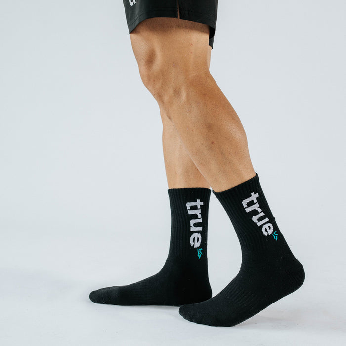 Black crew crew socks with white "true" logo on athletic male legs, product photo on white studio background — men's athleisure cotton-blend socks for running, training and everyday wear.