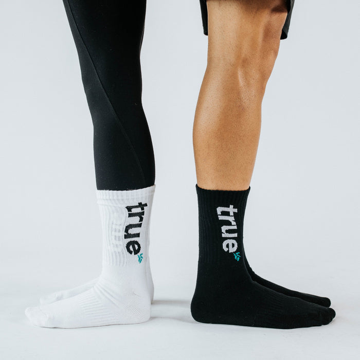 White and black unisex athletic crew socks with vertical "true" logo and teal leaf icon, modeled on two standing legs in studio on white background — product lifestyle shot for socks, activewear.