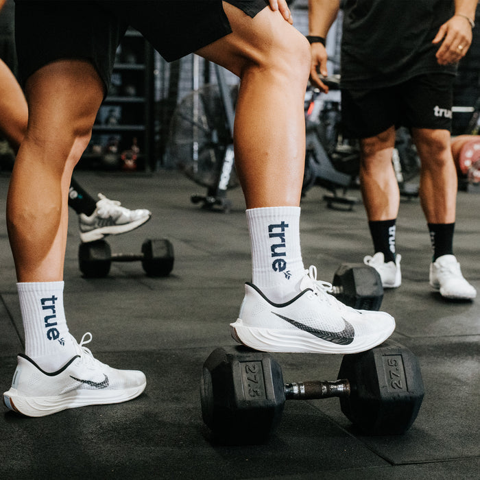 White crew athletic socks with navy "true" logo worn in gym on a model stepping on a 27.5 lb dumbbell, paired with white Nike running shoes in a group workout setting.